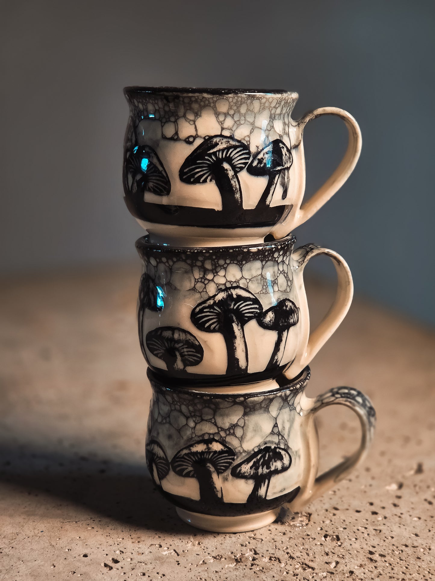 Three stacked off-white ceramic mushroom espresso mugs with black designs and a bubbly glaze, on a textured surface.