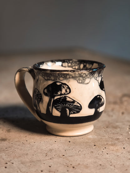 Off-white and black mushroom espresso mug with black mushroom designs and a mottled rim, sitting on a textured surface.