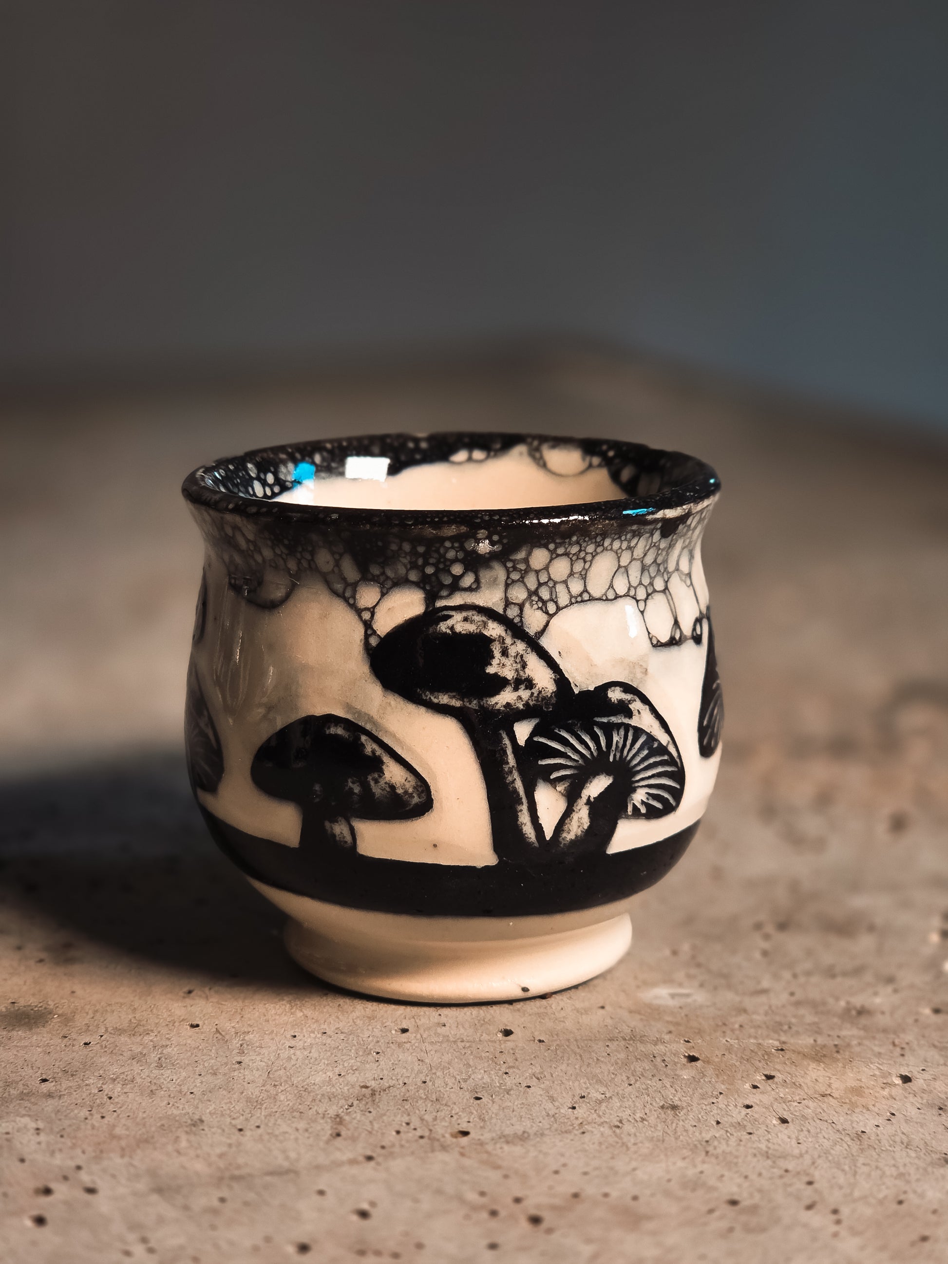Cream ceramic mushroom espresso mug featuring black mushroom illustrations and a bubbly glaze, on a textured surface.