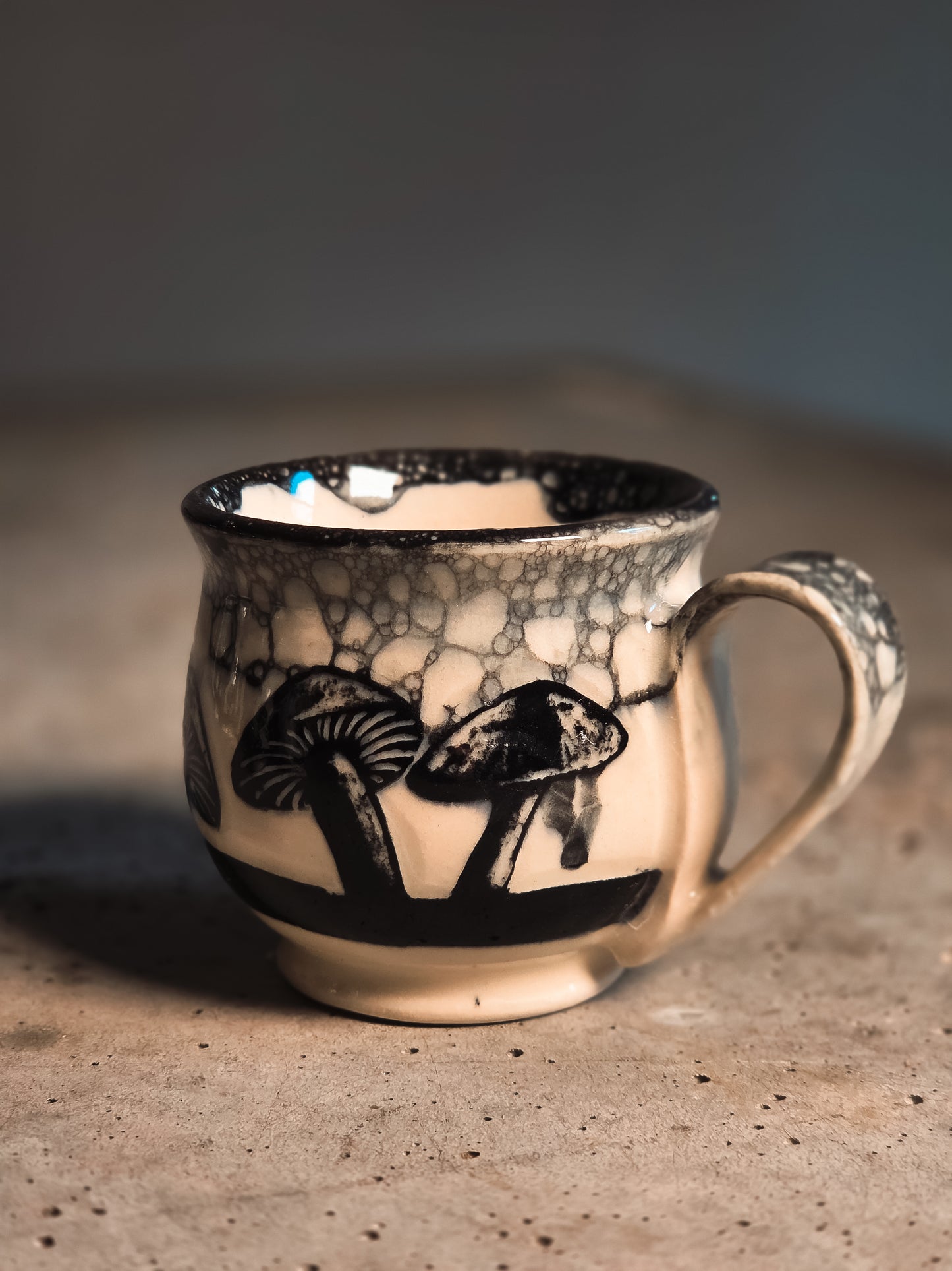 Cream espresso mug with dark mushroom illustrations, a bubble glaze rim, on a textured grey surface.