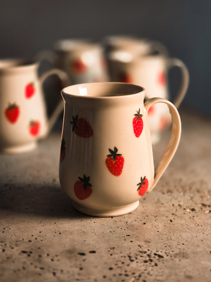 Cream ceramic strawberry mug with red fruit pattern, side view on speckled surface, multiple mugs in soft focus background