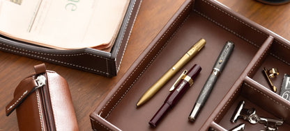 Brown leather supply tray with white stitching, holding various pens and stationery items on a wooden desk.