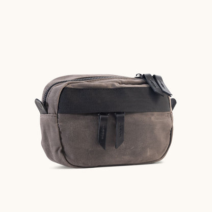 Slate grey waxed canvas crossbody mini bag with a top zipper, two black pull tabs, and a black front strap, angled.
