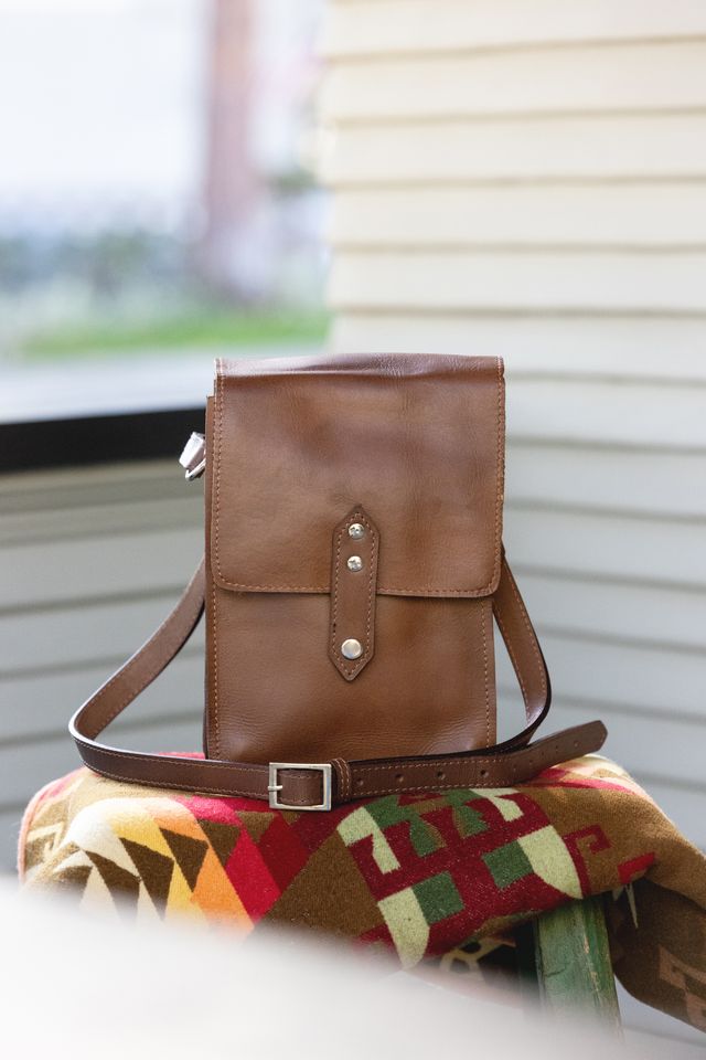 Brown leather crossbody bag with silver buckle and studs, resting on a colorful geometric patterned blanket.