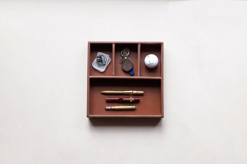 Brown leather supply tray with compartments holding three pens, keys, an SD card, and a golf ball.