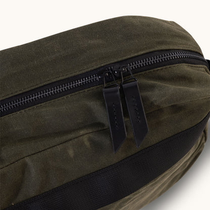 Juniper green waxed canvas crossbody bag with dark zippers, black leather pulls, and black webbing detail.