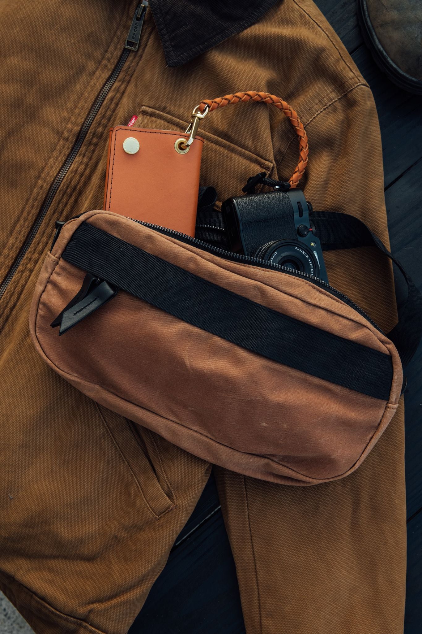 Desert Rose waxed canvas crossbody pack, unzipped, holding a tan leather wallet and black camera, on a brown canvas jacket.