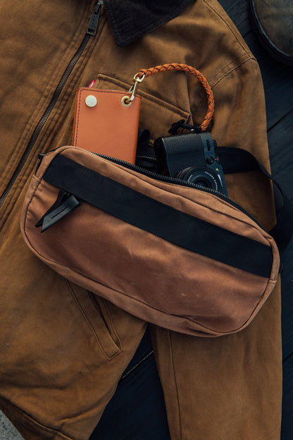 Desert Rose waxed canvas crossbody pack, unzipped, holding a tan leather wallet and black camera, on a brown canvas jacket.