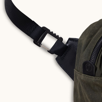Juniper green waxed canvas Canyon Crossbody bag, with a black webbing strap, black hook clip, zipper, and a small embossed leather logo tab.