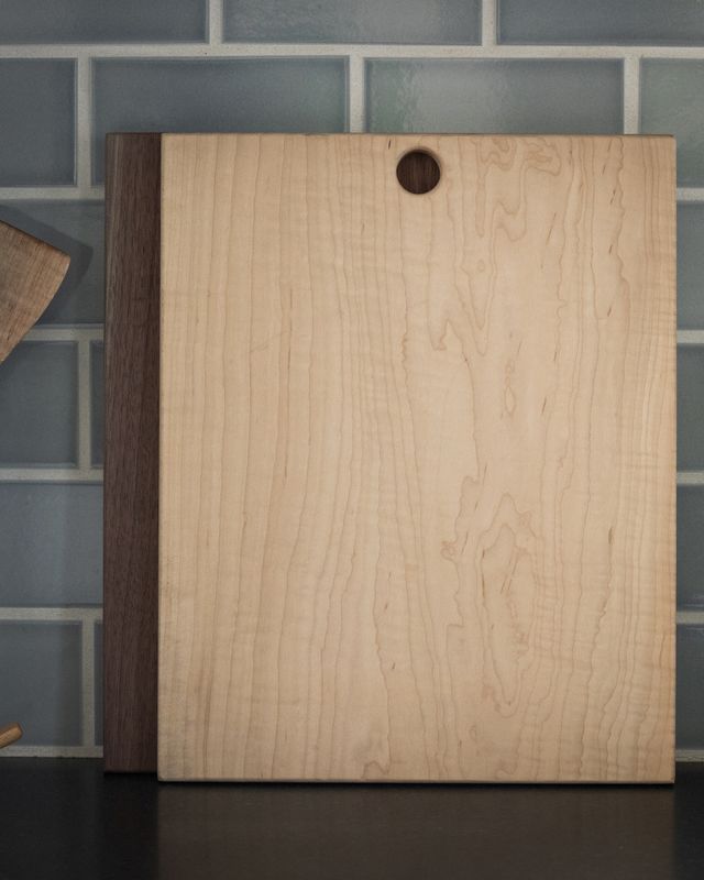 Light maple cutting board with a circular hole, leaning against blue subway tiles next to a darker wooden board.