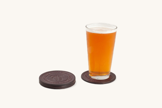 Dark brown round coaster set of 4 with an embossed logo, three stacked and one under a glass of beer.