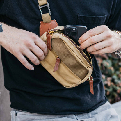 Tan crossbody mini bag with a visible zipper, brown leather pull tabs, and mesh front pocket, held by a person inserting a phone.