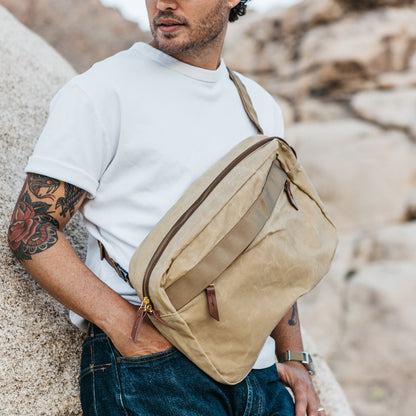 Tan waxed canvas Canyon Crossbody Large bag with dark brown zippers and leather pulls, worn by a man in a white t-shirt and jeans.