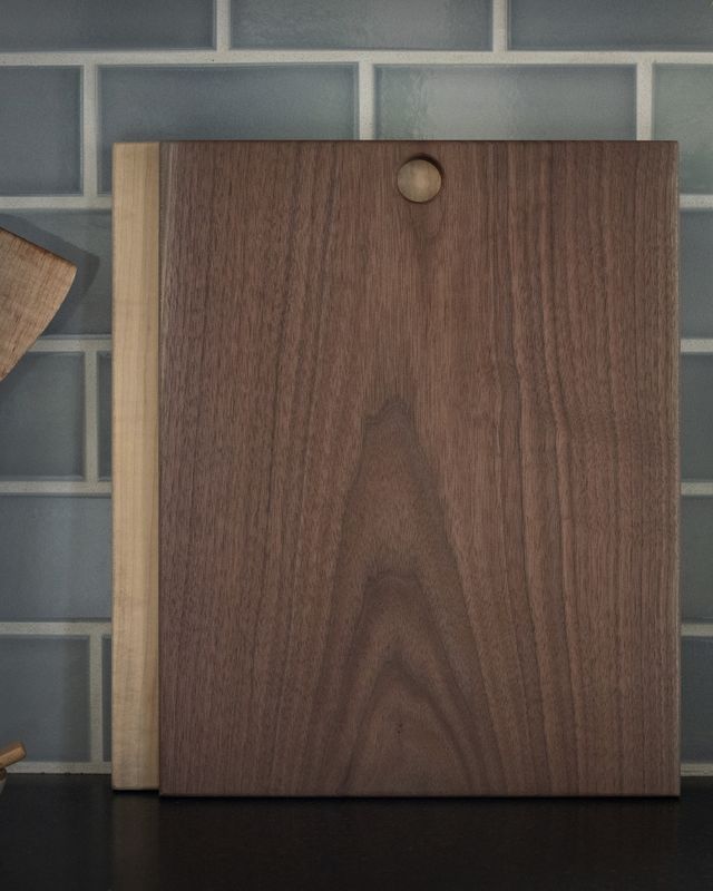 Walnut cutting board with a light wood strip, featuring a hanging hole, against a gray subway tile backsplash.