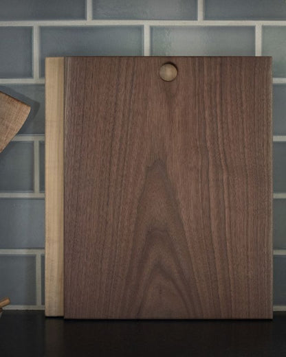 Walnut cutting board with a light wood strip, featuring a hanging hole, against a gray subway tile backsplash.