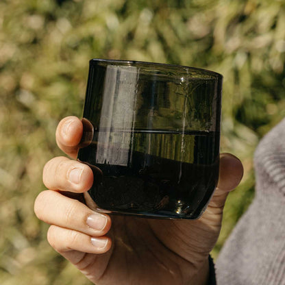 Dark green cocktail glass with liquid, held in a hand against a blurred green background.