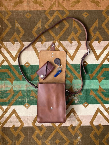 Brown leather crossbody bag, open to show a matching key case and keys, all on a geometric patterned rug.