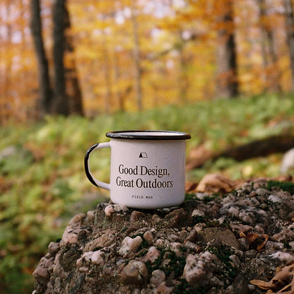 White enamel mug by Field Mag with 'Good Design, Great Outdoors' text and a tent icon, resting on a rock in an autumn forest.