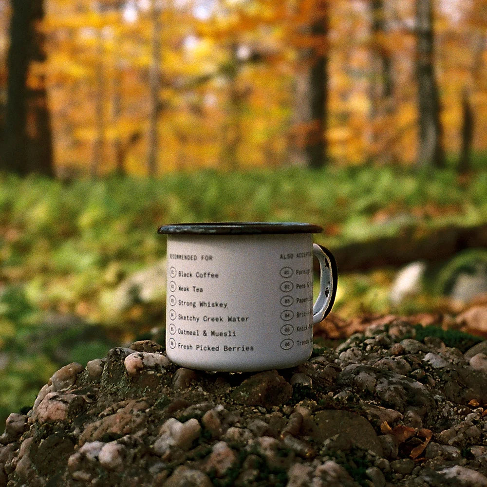 White enamel mug with a black rim by Field Mag, featuring humorous text lists, sitting on rocks in an autumn forest.