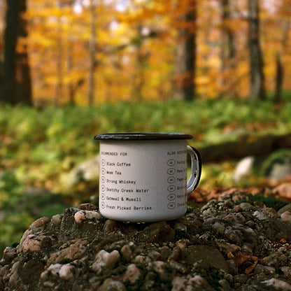 White enamel mug with a black rim by Field Mag, featuring humorous text lists, sitting on rocks in an autumn forest.