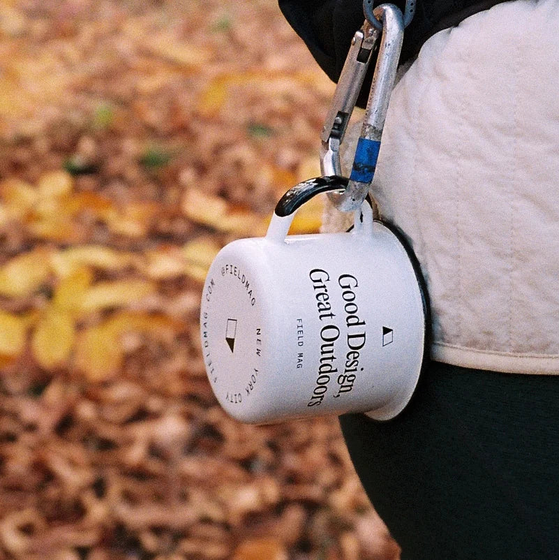 White enamel mug by Field Mag, printed with "Good Design, Great Outdoors," hanging from silver carabiners against blurred autumn leaves.