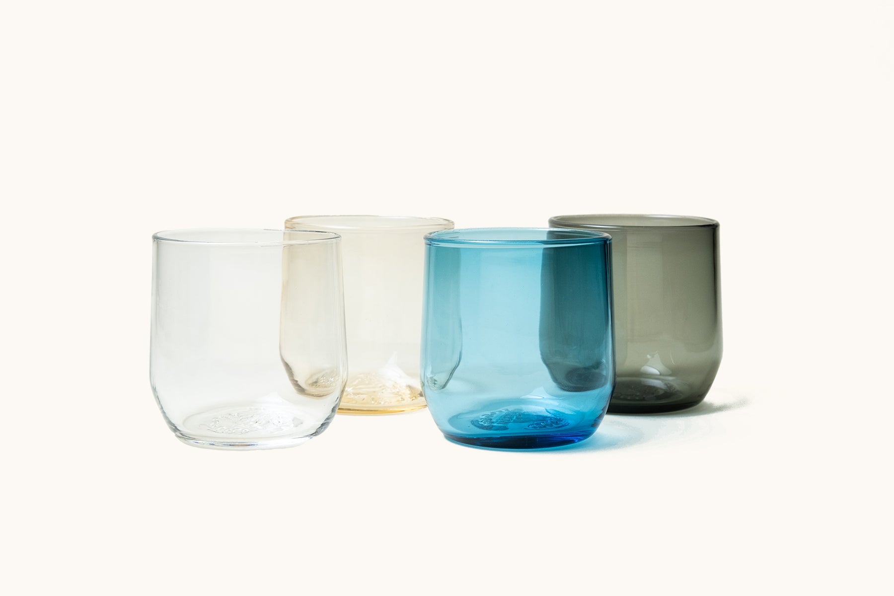 Four stemless cocktail glasses made of clear, amber, blue, and grey glass.