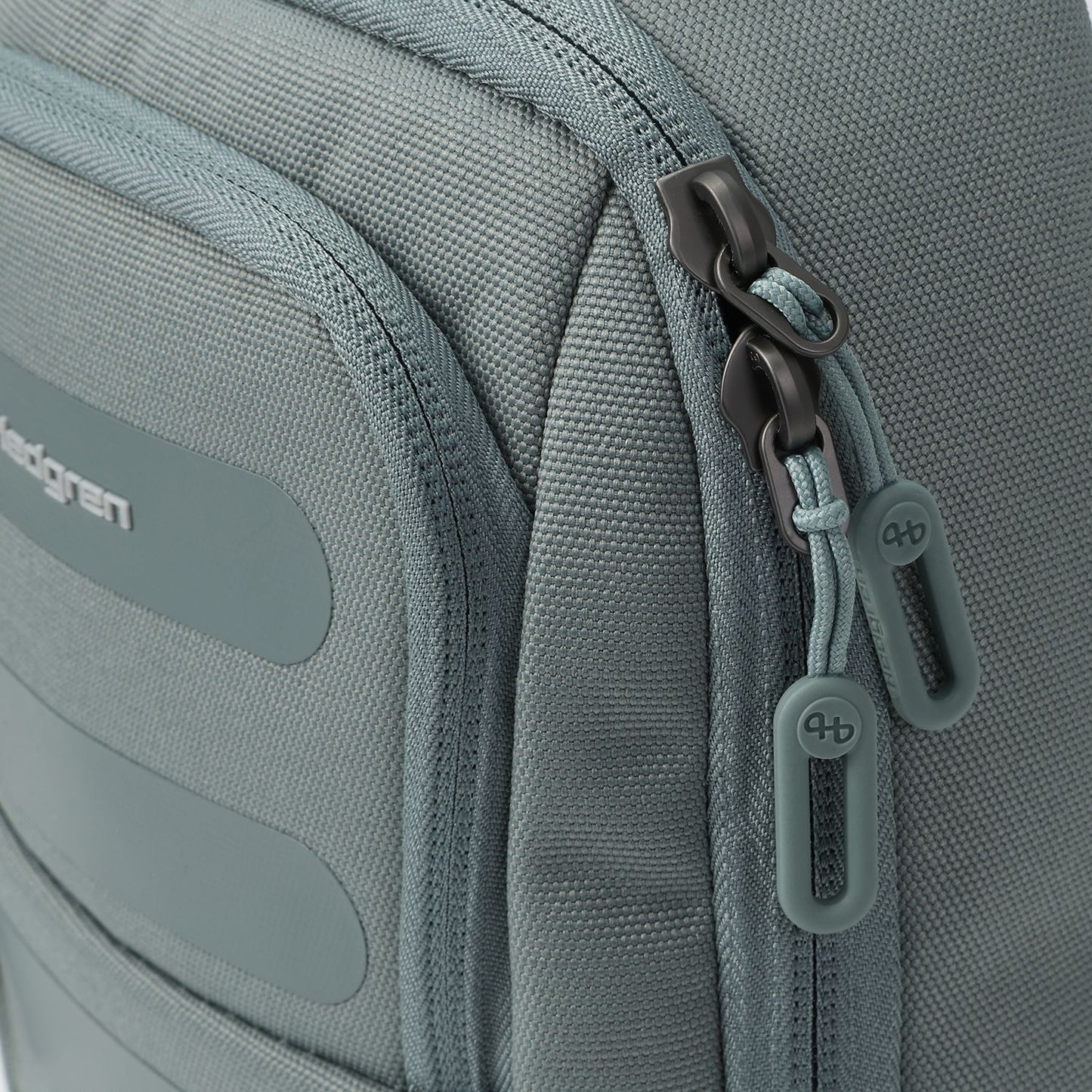 Muted grey sling bag by Hedgren with dark metal zippers, light teal cord loops, and rubber 'hb' logo pulls.