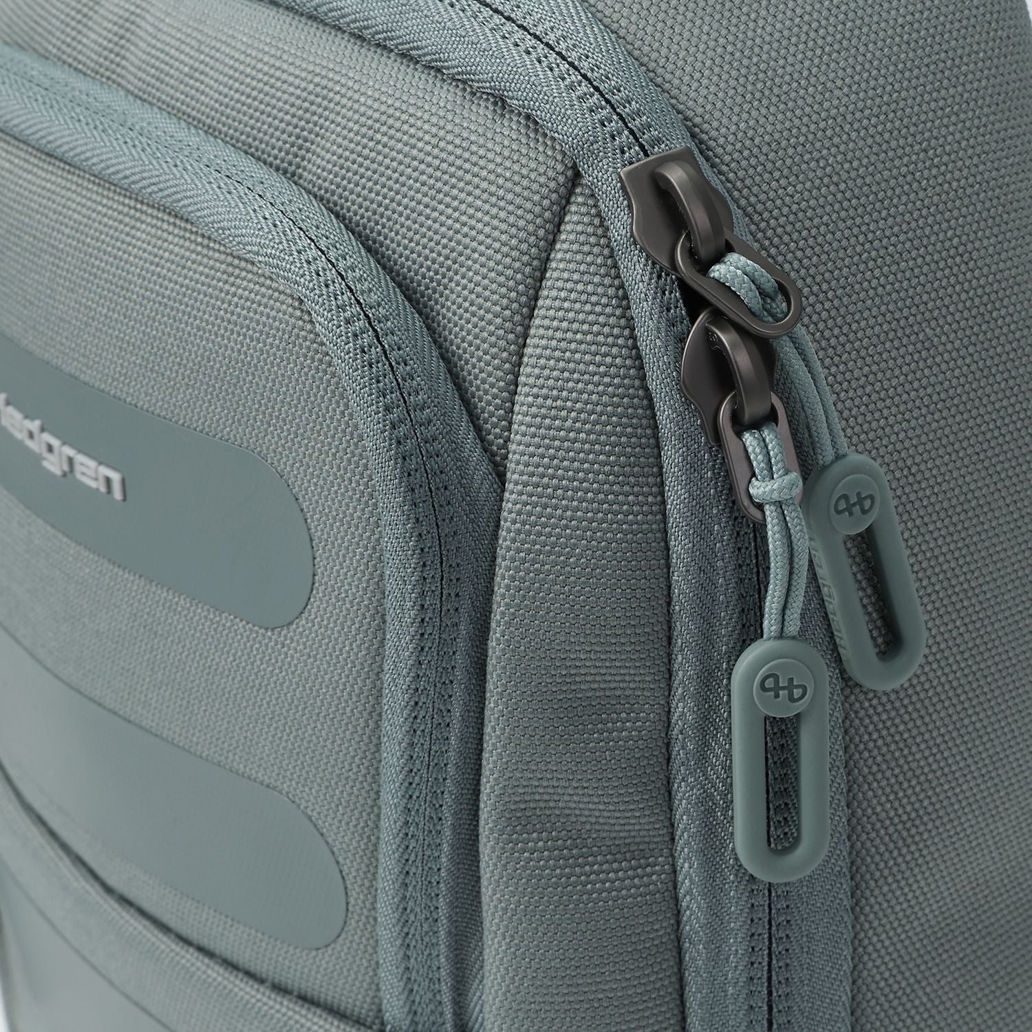 Muted grey sling bag by Hedgren with dark metal zippers, light teal cord loops, and rubber 'hb' logo pulls.