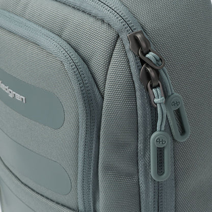 Muted grey sling bag by Hedgren with dark metal zippers, light teal cord loops, and rubber 'hb' logo pulls.