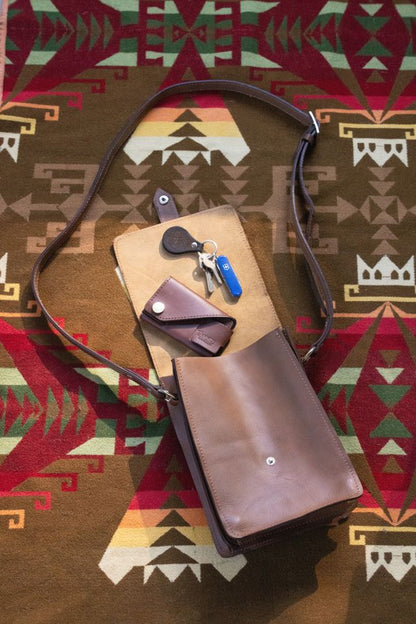 Brown leather crossbody bag, open to reveal keys and a small key holder, resting on a colorful patterned blanket.