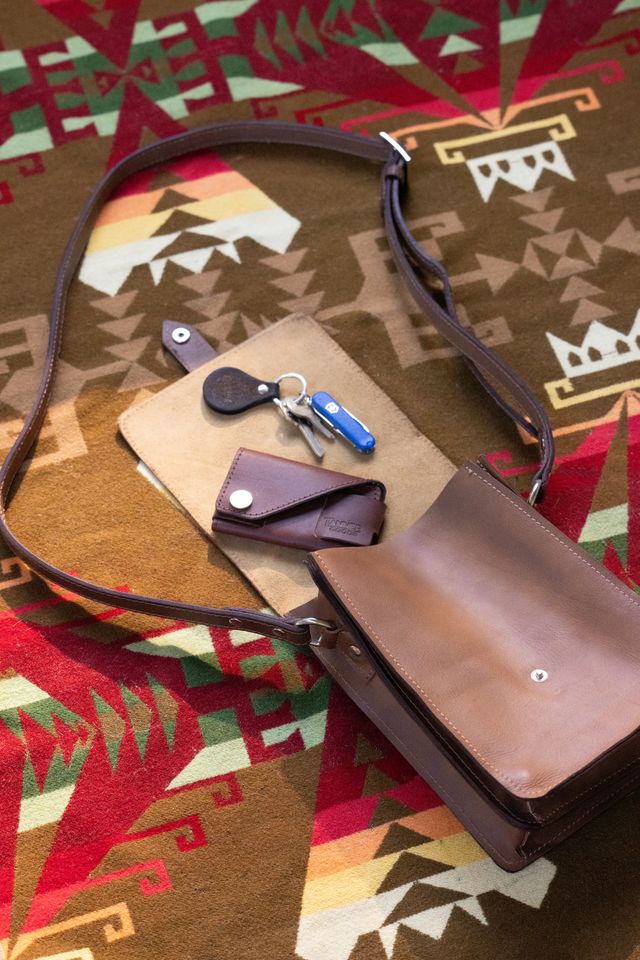 Brown leather crossbody bag with a flap closure, styled open on a colorful patterned blanket with keys and a small wallet.