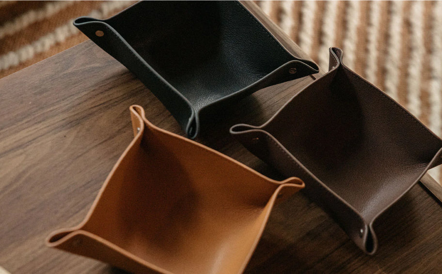 Three leather document trays in tan, brown, and black arranged on a dark wood table with striped rug background.