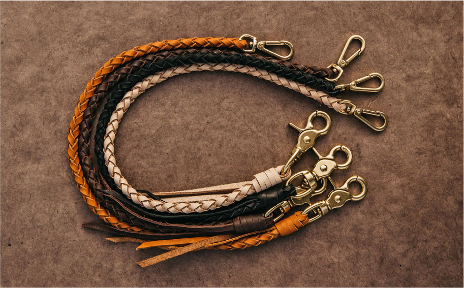 Four braided leather lanyards in orange, black, white, and brown with gold-tone swivel hooks, arranged in a loop on a textured brown surface.
