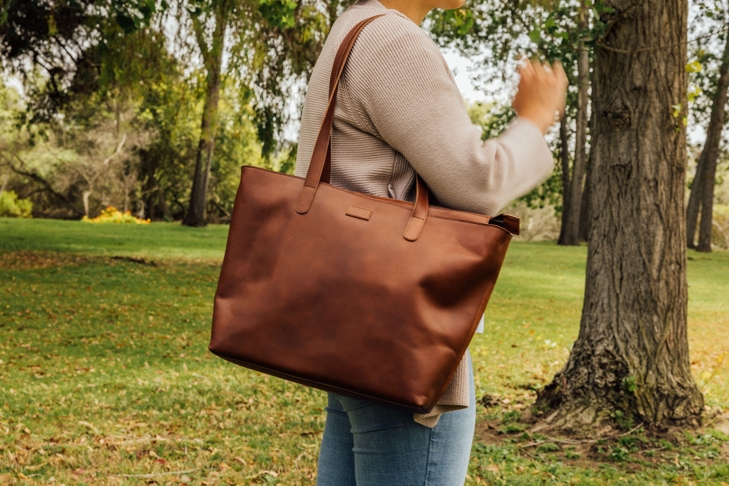 Woman in beige sweater and jeans carries large brown leather tote bag in sunlit park with green grass and trees.