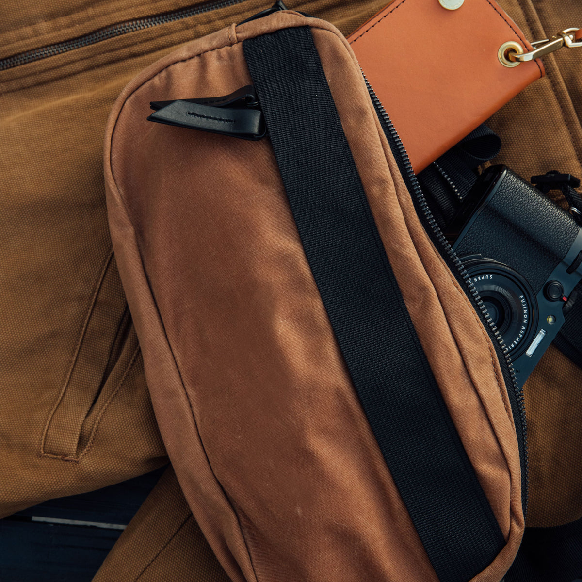 Brown waxed canvas Canyon Crossbody Pack with a black strap, partially unzipped, revealing a brown wallet and camera inside.