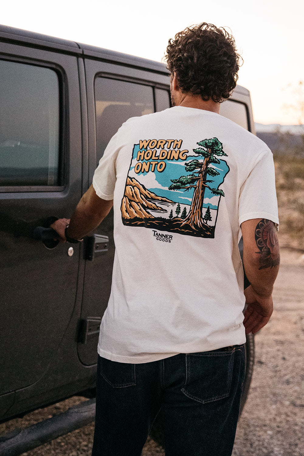 White Adventure T-Shirt from Tanner Goods featuring a scenic graphic and 'WORTH HOLDING ONTO' text on the back, worn by a person opening a car door.