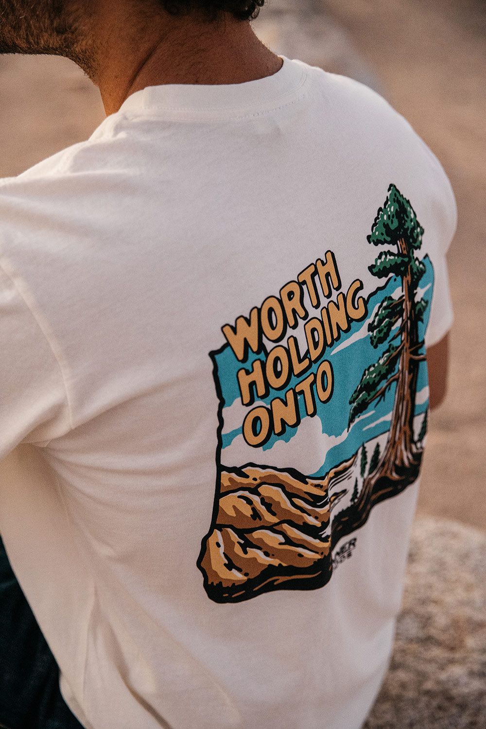 Cream t-shirt featuring a colorful "WORTH HOLDING ONTO" graphic of a tree and landscape on the back, worn by a person.
