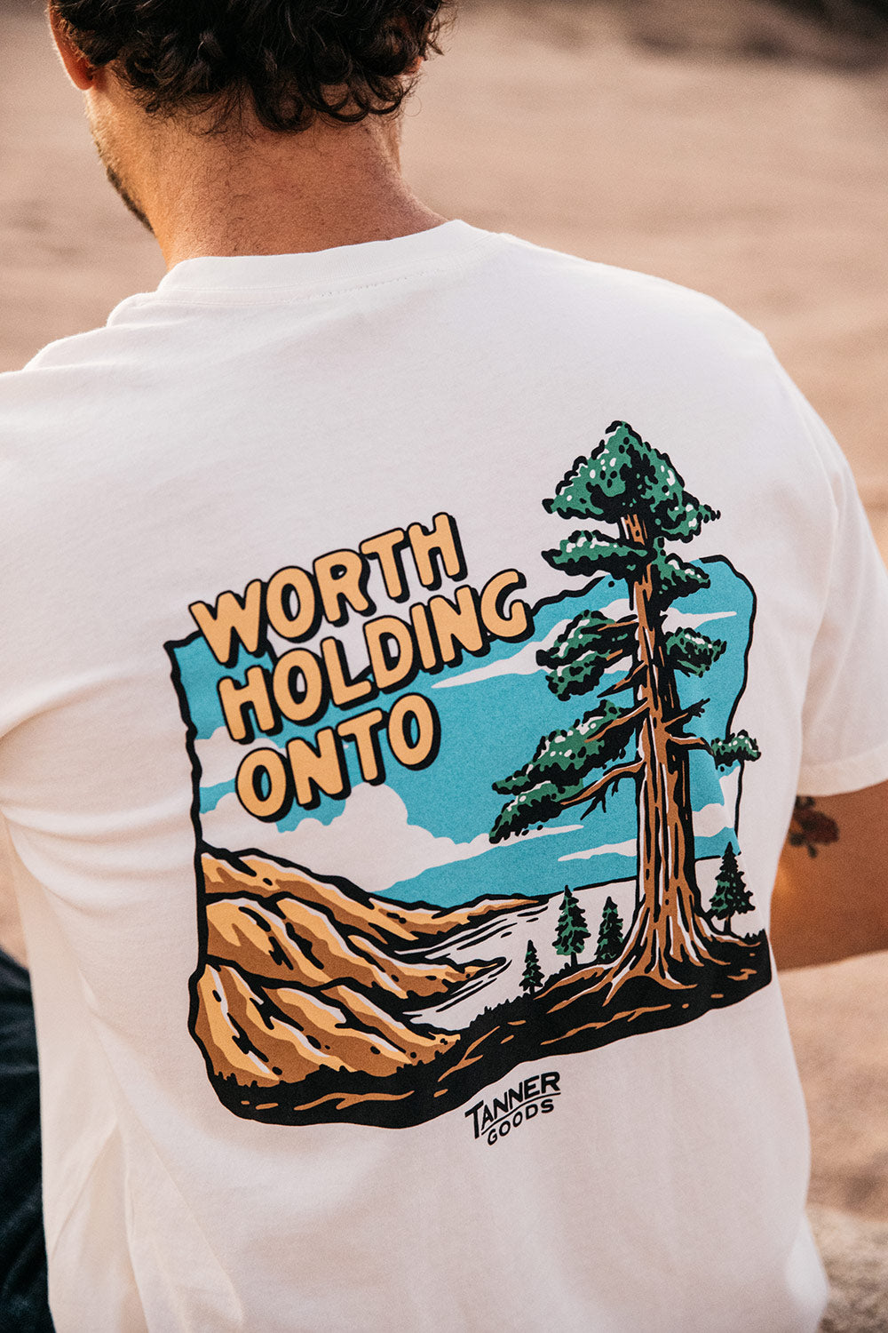White t-shirt by Tanner Goods, back view, featuring a nature graphic print with "Worth Holding Onto" text.