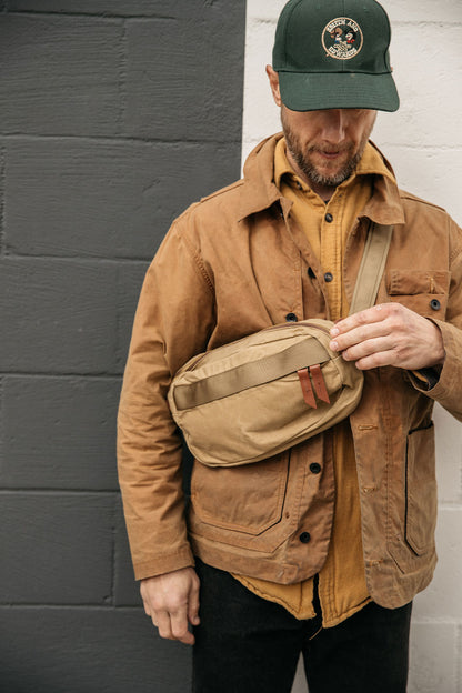 Utility Tan waxed canvas Canyon Crossbody Pack with brown leather zipper pull tabs, worn across a man's chest.