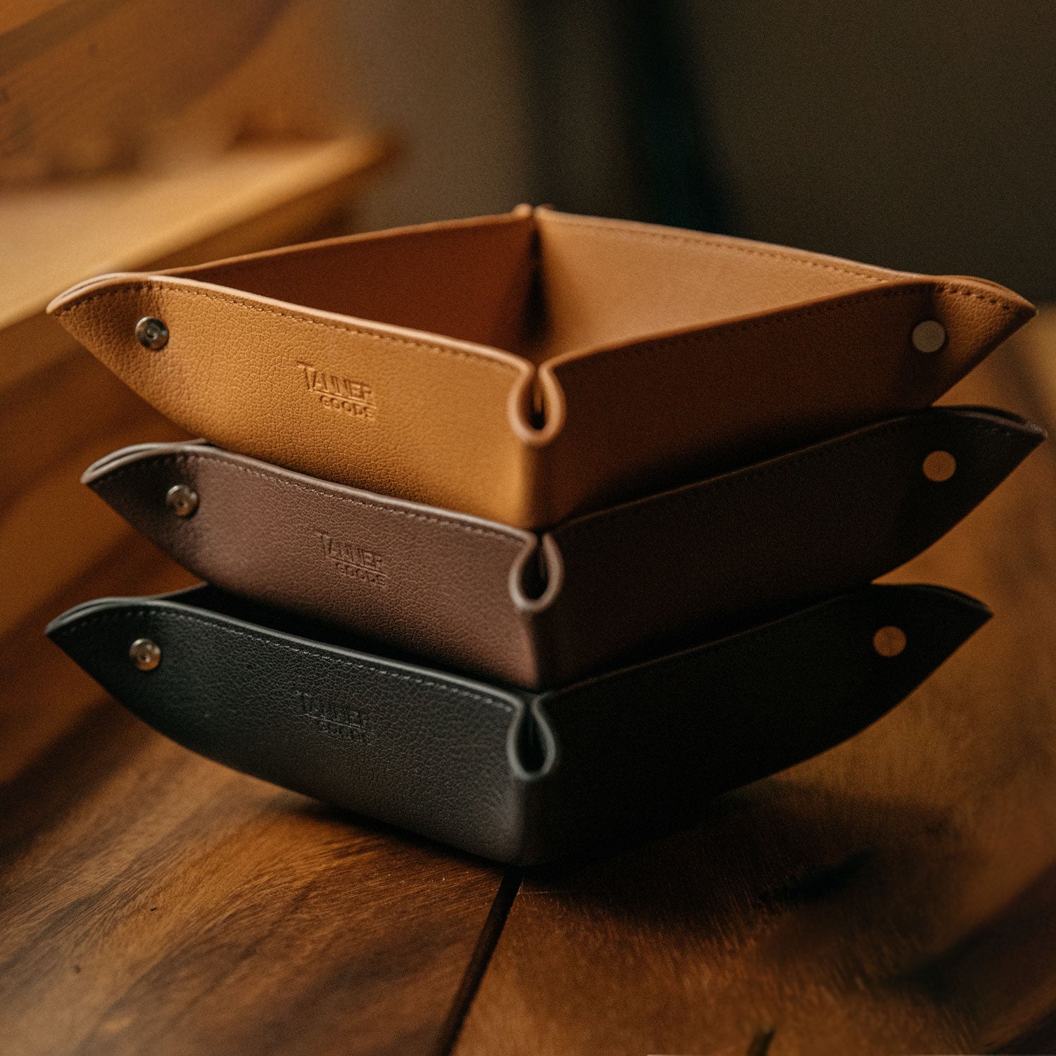 Black recycled leather valet tray, stacked on two other textured leather trays with metal snaps on a wooden surface.