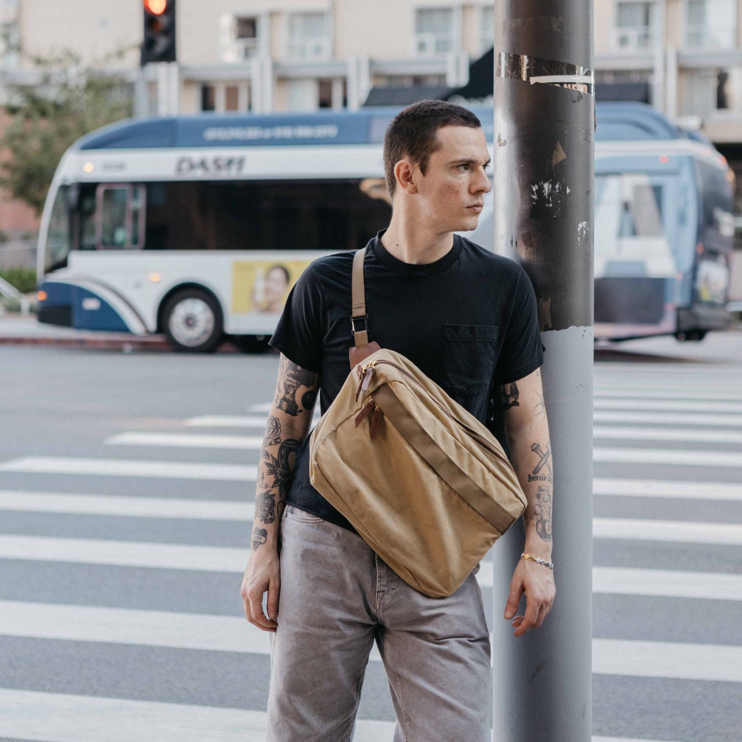 Tan canvas crossbody bag worn by a man with tattoos, leaning against a pole on a city street.