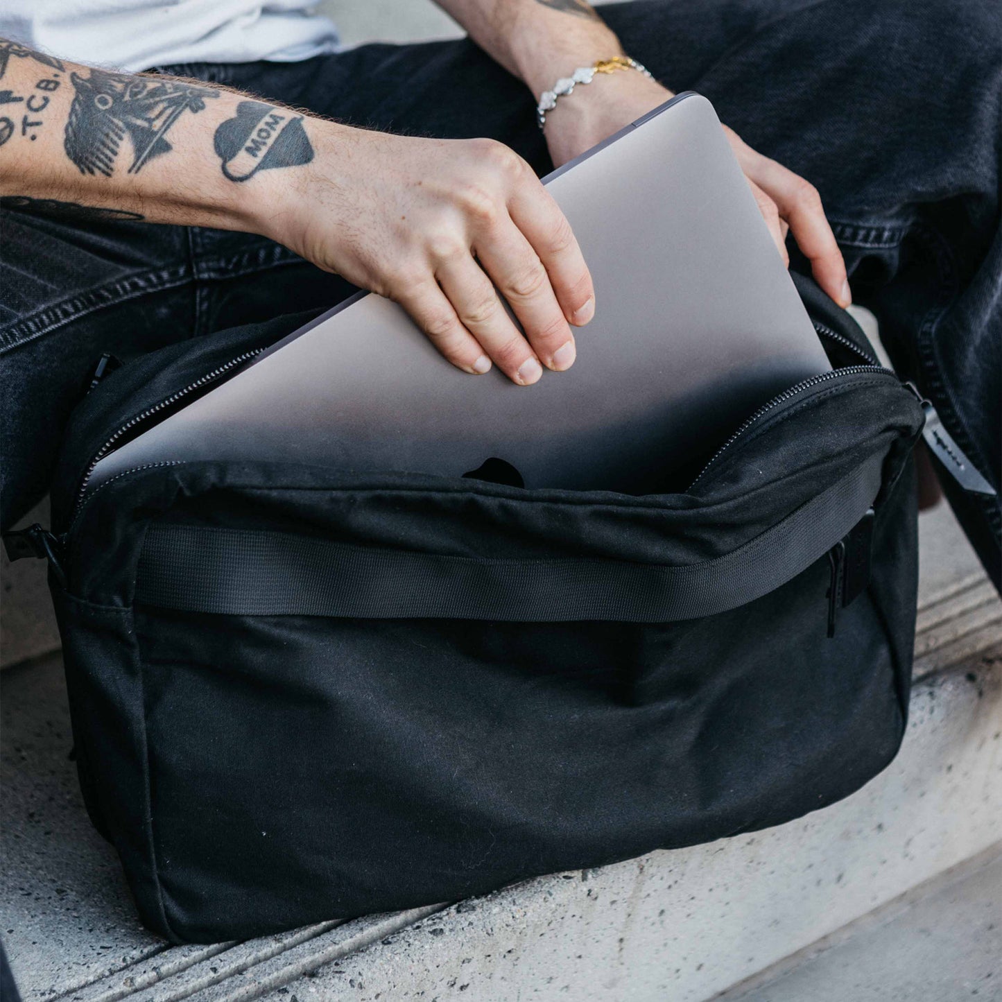 Large slate grey waxed canvas crossbody bag with a person inserting a laptop into the zippered top.