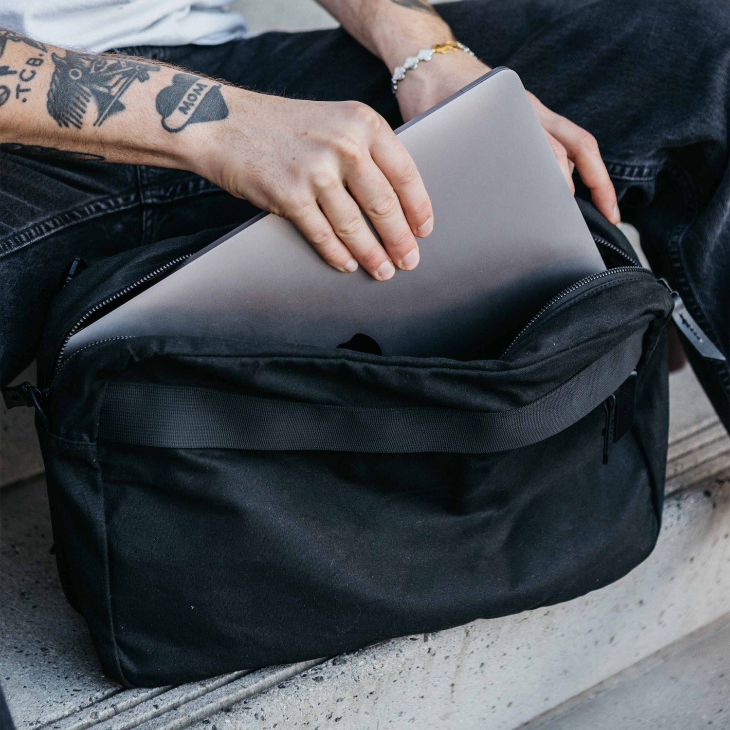 Large slate grey waxed canvas crossbody bag with a person inserting a laptop into the zippered top.