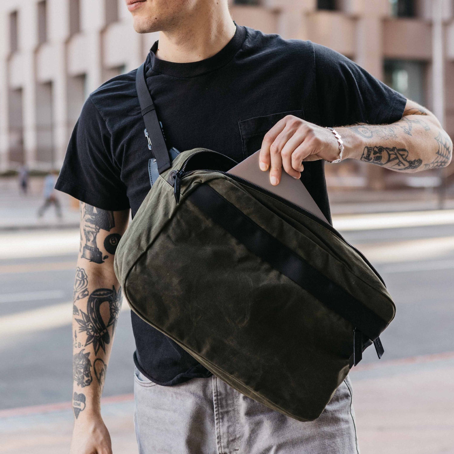 Canyon Crossbody Large in Slate Grey waxed canvas, carried by a man placing a silver laptop into its open main compartment.