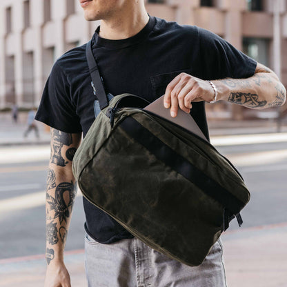 Canyon Crossbody Large in Slate Grey waxed canvas, carried by a man placing a silver laptop into its open main compartment.