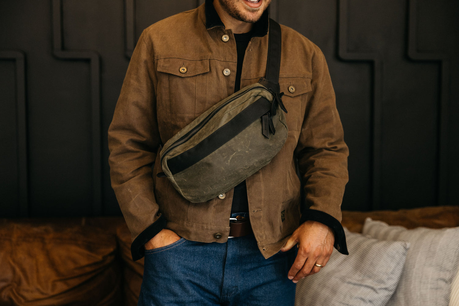 Man in brown jacket and blue jeans wears olive green crossbody bag with black strap, hands in pockets, standing against dark paneled wall.