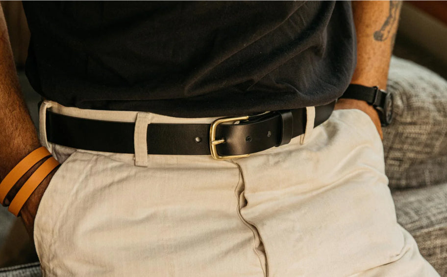 Close-up of person wearing black leather belt with gold buckle, beige pants, and black shirt, hands resting on thighs.
