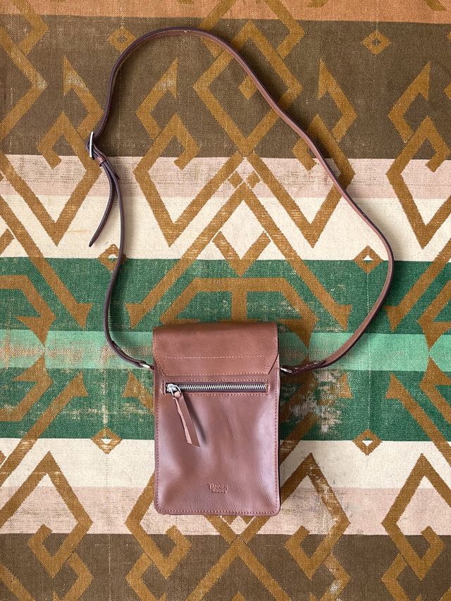 Tan leather crossbody bag with a front zipper pocket and flap closure, featuring an adjustable strap, on a geometric patterned rug.