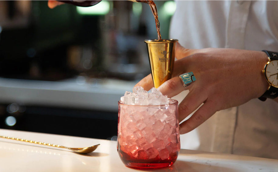 Barista pours dark liquid through gold jigger into pink glass filled with ice; hand wears turquoise ring, gold watch; spoon rests nearby on white counter.