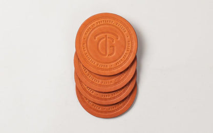 Four stacked round orange leather coasters, embossed with a TG logo and text, on a white background.